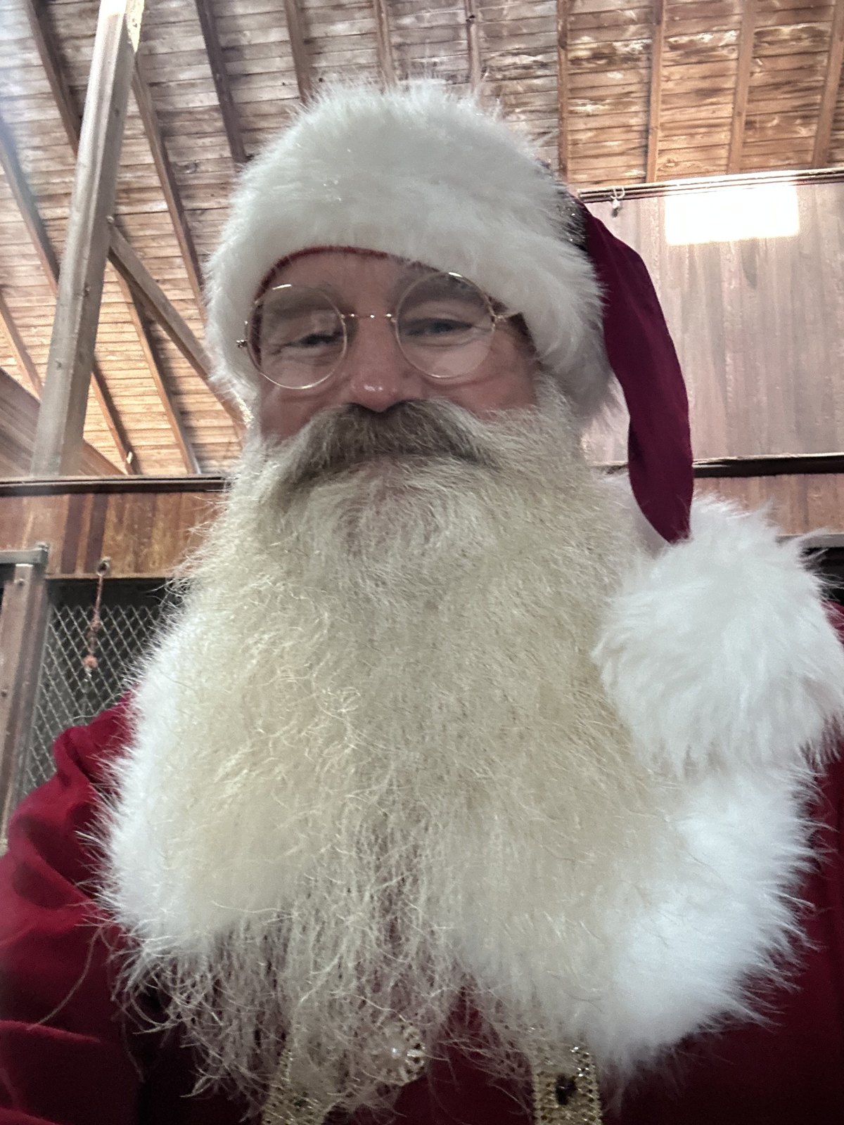 Santa smiling warmly inside a barn, wearing a deep red suit with white fur trim